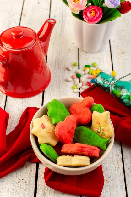 Vue de dessus de délicieux biscuits colorés différents formés à l'intérieur de la plaque avec des bonbons et des fleurs de bouilloire rouge