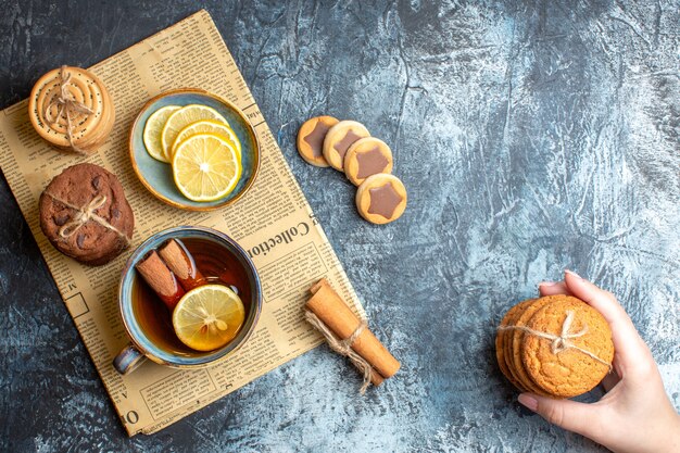 Vue de dessus de délicieux biscuits et main tenant une tasse de thé noir à la cannelle sur un vieux journal sur fond sombre