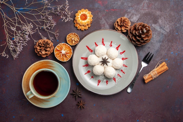 Vue de dessus de délicieux bonbons à la noix de coco petits et ronds formés avec une tasse de thé sur le fond sombre des bonbons à la noix de coco et des biscuits au gâteau sucré