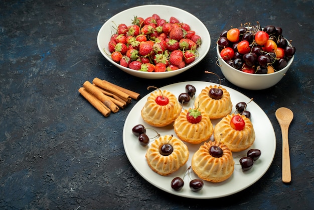 Une vue de dessus de délicieux cookies à l'intérieur de la plaque blanche avec des cerises fraises sur le gâteau aux fruits de bureau noir