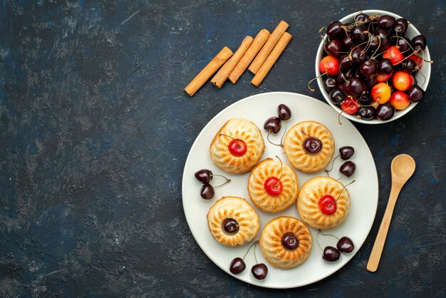Une vue de dessus de délicieux cookies à l'intérieur de la plaque blanche avec des cerises sur le gâteau aux fruits de bureau noir