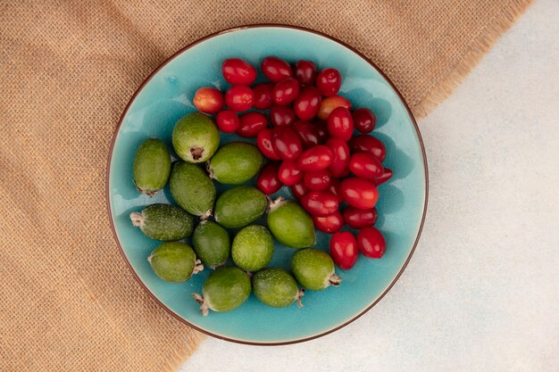 Vue de dessus de délicieux feijoas mûrs avec des cerises cornaline sur un plat bleu sur un tissu de sac sur une surface grise