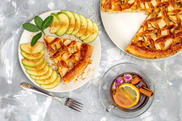 Une vue de dessus délicieux gâteau aux pommes à l'intérieur de la plaque avec du thé et de la pomme verte fraîche sur le sucre biscuit gâteau de bureau gris