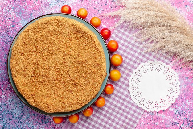 Vue de dessus délicieux gâteau rond à l'intérieur de la plaque avec des cerises-prunes bordées sur le bureau rose vif gâteau tarte biscuit sweet bake fruit