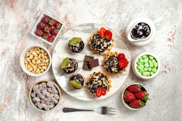 Vue de dessus de délicieux gâteaux crémeux avec des baies et des bonbons sur un sol blanc biscuit gâteau aux fruits tarte à la crème biscuit au thé