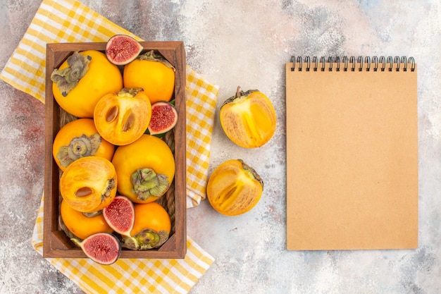 Vue de dessus de délicieux kakis et figues coupées dans une boîte en bois serviette de cuisine jaune un cahier sur fond nu