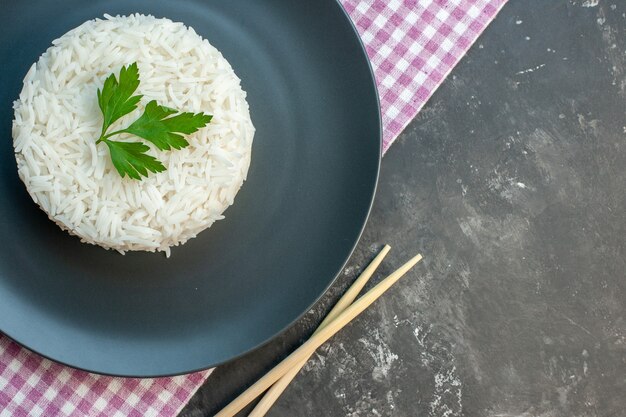 Vue de dessus d'un délicieux repas de riz servi avec du vert sur une plaque noire sur une serviette à rayures violettes et des baguettes en bois sur le côté droit sur fond sombre