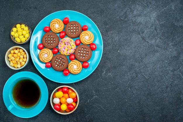 Vue de dessus différents biscuits au sucre avec des bonbons et une tasse de thé sur le bureau gris bonbons biscuits au thé sucré