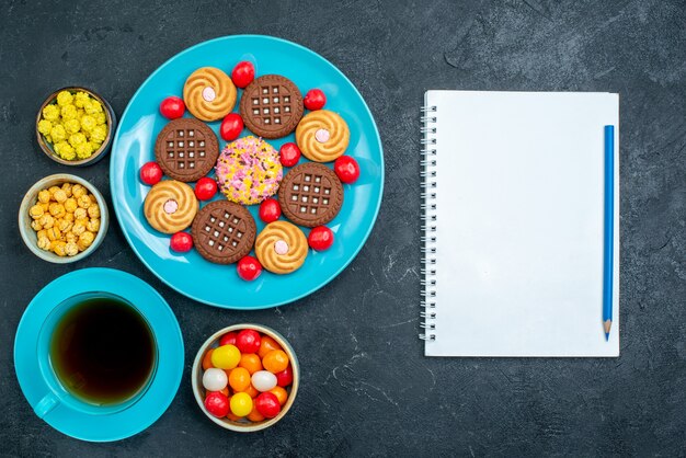 Vue de dessus différents biscuits au sucre avec des bonbons et une tasse de thé sur la surface grise sucre candy biscuit thé sucré biscuit