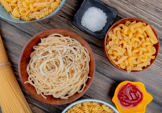 Vue de dessus de différents macaronis comme spaghetti rotini vermicelles et autres avec du sel et du ketchup sur bois