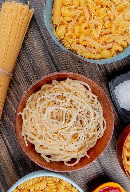 Vue de dessus de différents types de pâtes comme les spaghetti rotini vermicelles et autres avec du sel et du ketchup sur une surface en bois