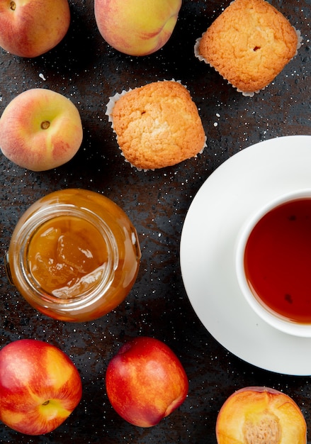 Photo gratuite vue de dessus du bocal en verre de confiture de pêches avec des cupcakes aux pêches et une tasse de thé sur une surface noire et brune