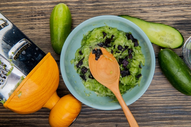 Vue de dessus du bol de salade de légumes avec une cuillère en bois et des concombres avec une râpe sur une surface en bois