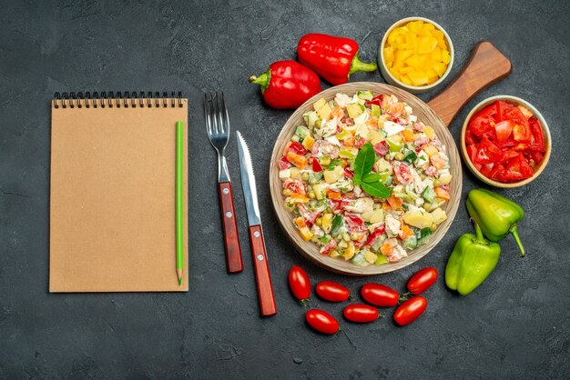 Vue de dessus du bol de salade de légumes avec des légumes couverts et bloc-notes sur le côté sur la table gris foncé