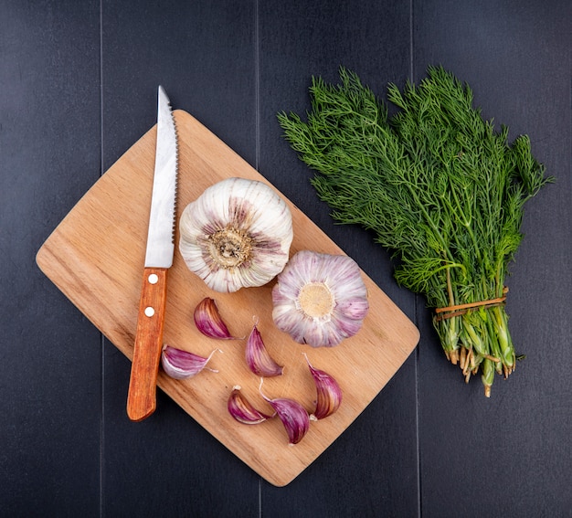 Photo gratuite vue de dessus du bulbe d'ail et des gousses avec un couteau sur une planche à découper et un bouquet d'aneth sur une surface noire