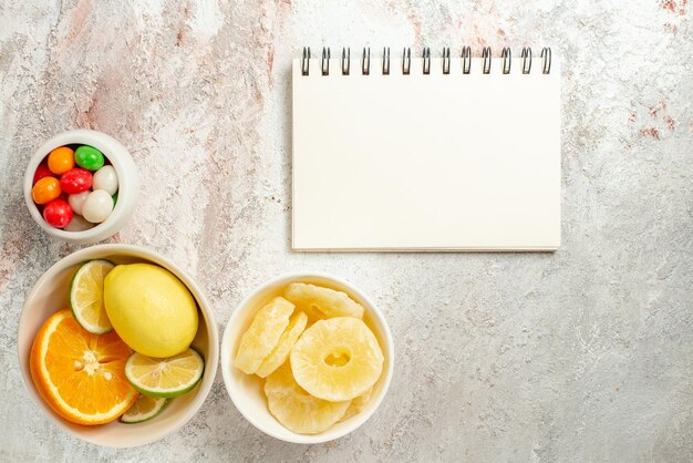 Vue de dessus du cahier blanc d'agrumes à côté des bols de bonbons colorés, d'ananas séchés et d'agrumes sur la table