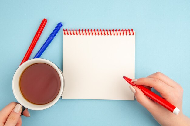 Vue de dessus du cahier à spirale tasse de thé et marqueur rouge dans les mains des femmes marqueurs rouges et bleus sur bleu