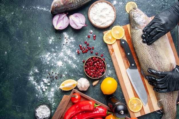 Vue de dessus du chef avec des gants noirs tenant du poisson cru sur une planche de bois moulin à poivre bol de farine graines de grenade dans un bol sur la table de la cuisine