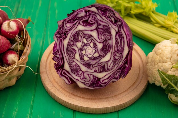 Vue de dessus du chou violet frais sur une planche de cuisine en bois avec des radis sur un seau avec chou-fleur et céleri isolé sur un mur en bois vert