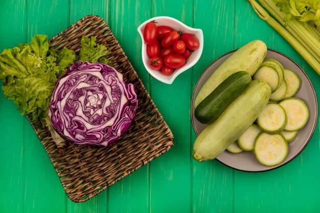 Vue de dessus du chou violet sur un plateau en osier avec de la laitue aux courgettes et concombre sur une assiette avec des tomates italiennes sur un bol de céleri isolé sur un mur en bois vert