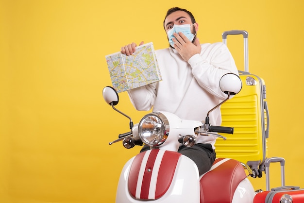 Vue de dessus du concept de voyage avec un gars confus dans un masque médical debout près d'une moto avec une valise jaune dessus et tenant une carte