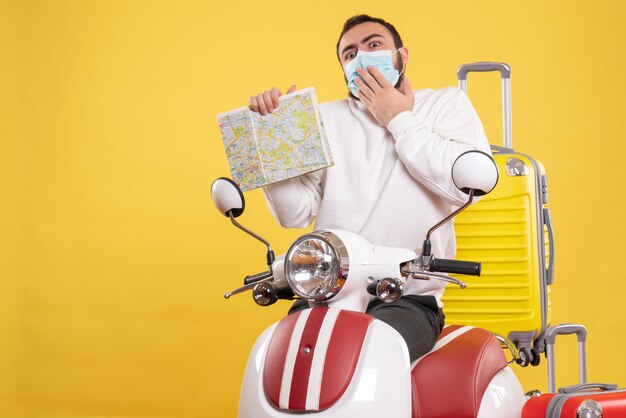 Vue de dessus du concept de voyage avec un gars confus dans un masque médical debout près d'une moto avec une valise jaune dessus et tenant une carte