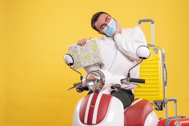 Vue de dessus du concept de voyage avec un gars rêveur dans un masque médical debout près d'une moto avec une valise jaune dessus et tenant une carte