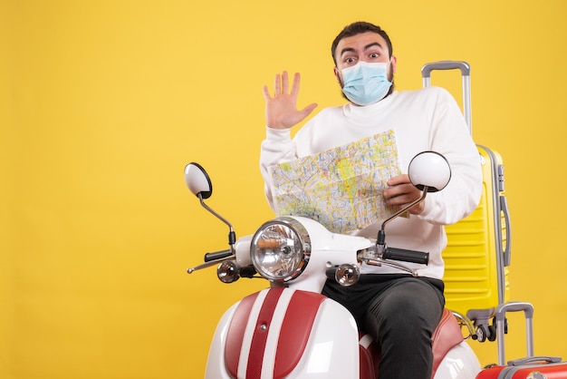 Vue de dessus du concept de voyage avec un gars surpris en masque médical assis sur une moto avec une valise jaune dessus et tenant une carte