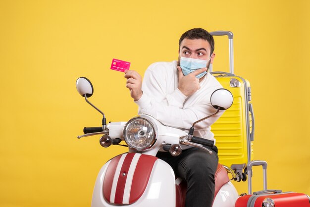 Vue de dessus du concept de voyage avec un jeune homme concentré en masque médical assis sur une moto avec une valise jaune dessus et tenant une carte bancaire