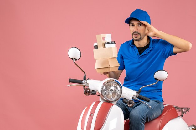 Vue de dessus du courrier homme portant un chapeau assis sur un scooter montrant les commandes se sentant confuses sur fond de pêche pastel