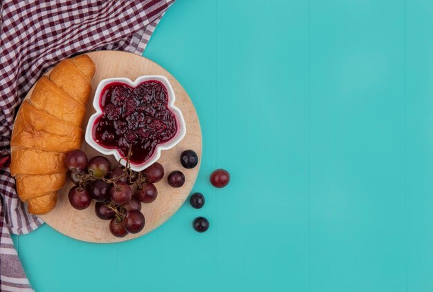 Vue de dessus du croissant et de la confiture de framboises dans un bol de raisin sur une planche à découper sur un tissu à carreaux sur fond bleu avec copie espace