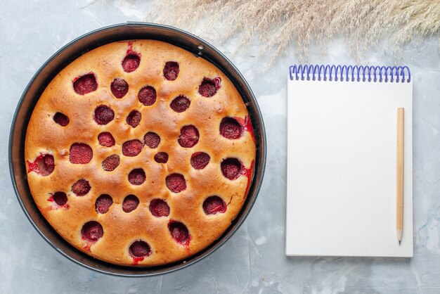 Vue de dessus du délicieux gâteau aux fraises cuit au four avec des fraises rouges fraîches à l'intérieur avec casserole et bloc-notes sur blanc, gâteau biscuit aux fruits