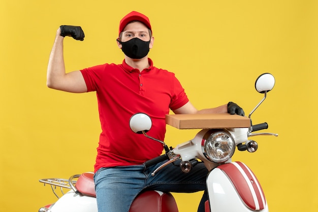 Vue de dessus du fier homme de messagerie ambitieux portant chemisier rouge et gants de chapeau en masque médical assis sur un scooter montrant l'ordre