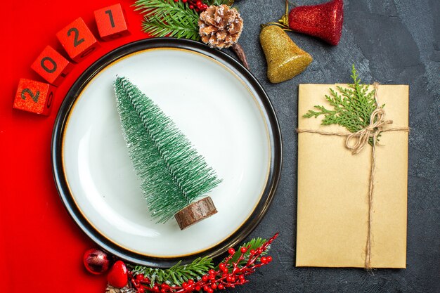 Vue de dessus du fond de nouvel an avec arbre de Noël sur assiette à dîner accessoires de décoration branches de sapin et numéros sur une serviette rouge à côté de cadeau sur une table noire