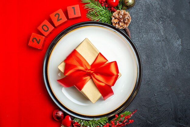 Vue de dessus du fond de nouvel an avec cadeau sur assiette à dîner accessoires de décoration branches de sapin et numéros sur une serviette rouge sur un tableau noir