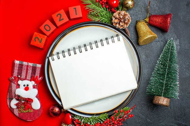 Vue de dessus du fond de nouvel an avec cahier sur assiette à dîner accessoires de décoration branches de sapin et numéros sur une serviette rouge à côté de l'arbre de Noël sur une table noire