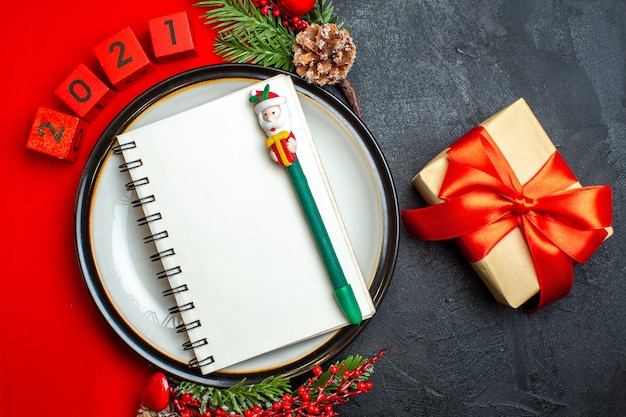 Vue de dessus du fond de nouvel an avec cahier à spirale sur assiette à dîner accessoires de décoration branches de sapin et numéros sur une serviette rouge et cadeau avec ruban rouge sur une table noire