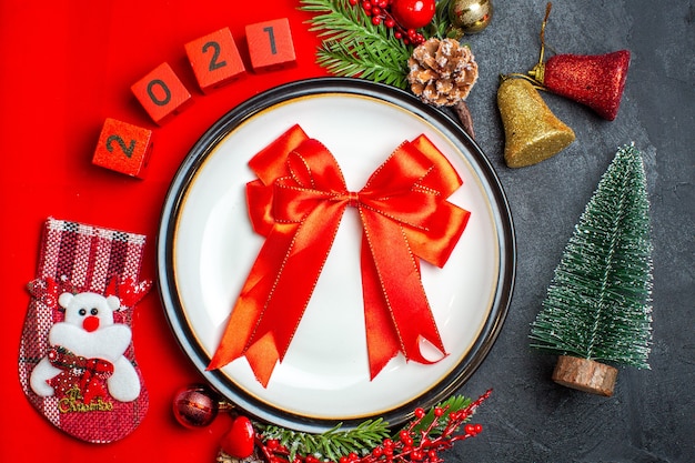 Vue de dessus du fond de nouvel an avec ruban rouge sur assiette à dîner accessoires de décoration branches de sapin et numéros chaussette de Noël sur une serviette rouge à côté de l'arbre de Noël sur une table noire