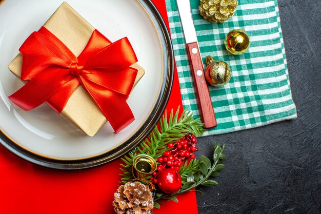Vue de dessus du fond de repas de Noël national avec cadeau avec ruban rouge en forme d'arc sur des assiettes vides couverts accessoires de décoration sur une serviette dépouillé vert