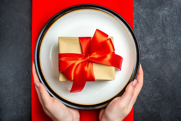 Vue de dessus du fond de repas de Noël national avec main tenant des assiettes vides avec ruban rouge en forme d'arc sur une serviette rouge sur le tableau noir