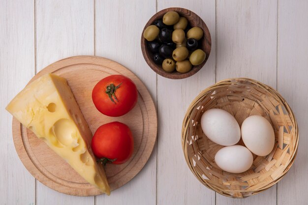 Vue de dessus du fromage maasdam aux tomates sur un support avec des olives et des œufs de poule dans un panier sur fond blanc