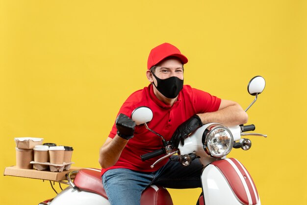 Vue de dessus du gars de courrier émotionnel confiant portant chemisier rouge et gants de chapeau dans un masque médical délivrant la commande assis sur un scooter