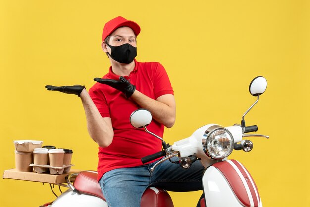 Vue de dessus du gars de courrier émotionnel portant un chemisier rouge et des gants de chapeau dans un masque médical délivrant une commande assis sur un scooter se sentant curieux