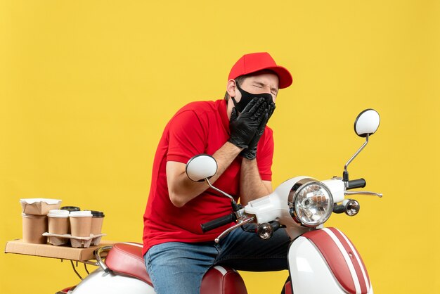 Vue de dessus du gars de messagerie en difficulté portant un chemisier rouge et des gants de chapeau dans un masque médical délivrant l'ordre assis sur un scooter souffrant de maux de dents