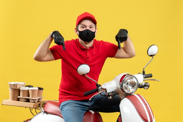 Vue de dessus du gars de messagerie portant un chemisier rouge et des gants de chapeau dans un masque médical délivrant la commande assis sur un scooter pointant vers le bas