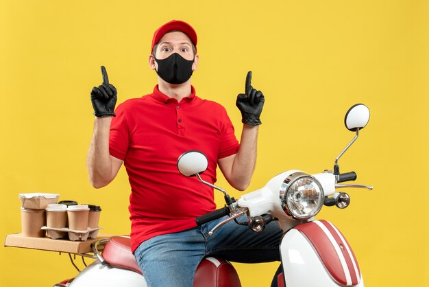 Vue de dessus du gars de messagerie surpris portant chemisier rouge et gants de chapeau en masque médical délivrant l'ordre assis sur un scooter pointant vers le haut