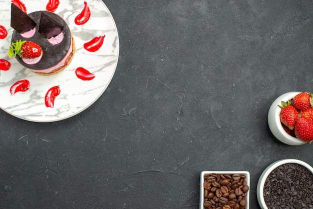 Vue de dessus du gâteau au fromage aux fraises sur une assiette ovale blanche en haut à gauche et des bols avec des graines de café au chocolat aux fraises en bas à droite de la surface sombre