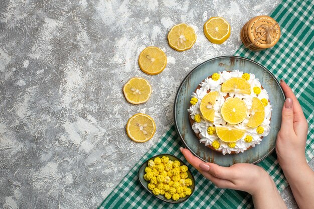 Vue de dessus du gâteau avec de la crème pâtissière et du citron sur un plateau dans des bonbons de biscuits à la main féminine dans un bol sur une nappe à carreaux blanc vert