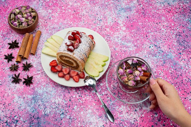 Vue de dessus du gâteau de rouleau à l'intérieur de la plaque avec des pommes et des fraises avec de la cannelle et du thé sur le gâteau de bureau de couleur biscuit fruit sucré