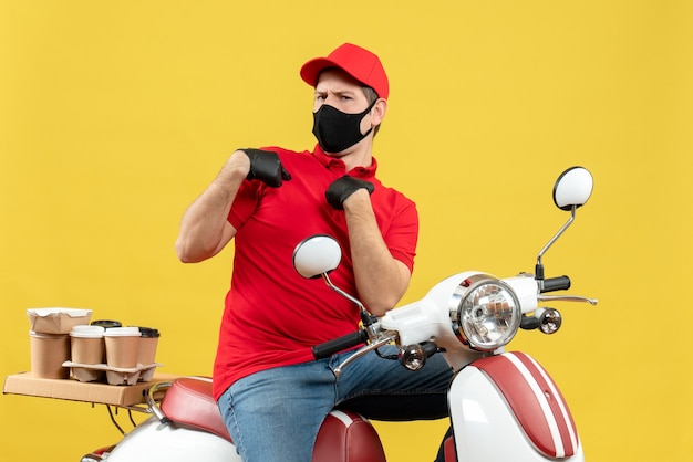 Vue de dessus du jeune adulte incertain incertain portant un chemisier rouge et des gants de chapeau dans un masque médical délivrant l'ordre assis sur un scooter sur un mur jaune